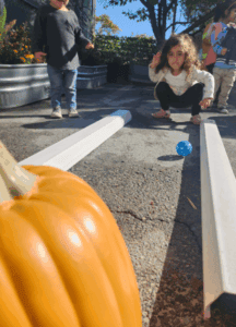 Toddler Engaging In A Homemade Bowling Game, Where A Ball Rolls Down A Self-Crafted Lane Towards A Pumpkin.
